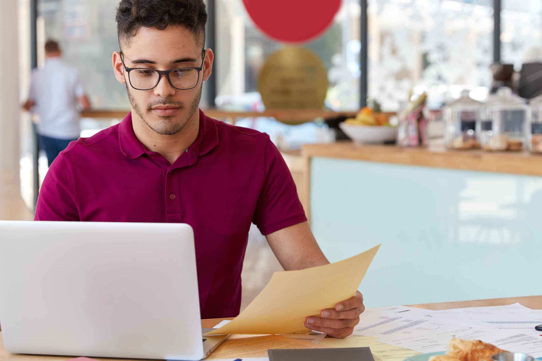 confident-man-casual-outfit-provides-electronic-payment-uses-online-banking-application-laptop-computer-develops-website-page-holds-papers-works-cozy-coffee-shop-modern-technologies